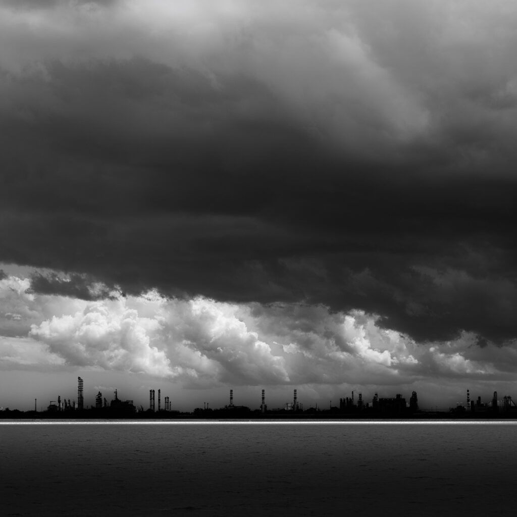 Ripresa dal mare, la zona industriale si mostra con le sue linee dure e con le sue torri sotto un cielo carico di nubi. Il forte contrasto ne esalta le geometrie severe, mentre una linea di luce proiettata sul mare sembra allentare la tensione poetica